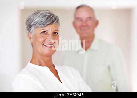 Lächelnde reife Frau mit Mann im Hintergrund. Porträt einer selbstbewussten, reifen Frau, die mit einem gutaussehenden Mann im Hintergrund lächelt. Stockfoto