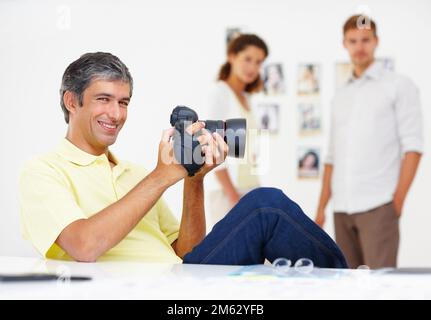 Kluger Mann, der Bilder in der Kamera überprüft. Porträt eines cleveren Mannes, der Fotos in seiner Kamera mit Kollegen im Hintergrund ansieht. Stockfoto