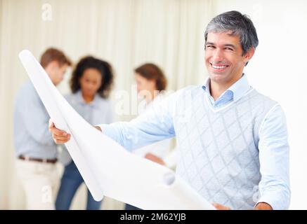 Ein Geschäftsmann, der den Plan durchsucht. Lächelnder Geschäftsmann liest Blaupausen mit Kollegen, die im Hintergrund diskutieren. Stockfoto