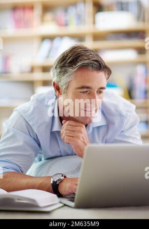 So viel, dass man sich nie langweilen kann. Ein gut aussehender, reifer Mann, der mit einem Laptop auf dem Boden seines Wohnzimmers liegt. Stockfoto
