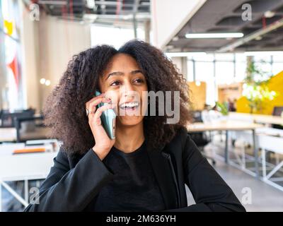 Positive junge afroamerikanische Unternehmerin in formellen Outfits, die am Tisch sitzt und telefoniert Stockfoto