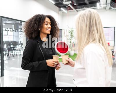 Frauen mit positiver ethnischer Herkunft in stilvoller Kleidung stehen in der Pause am modernen Arbeitsplatz und genießen aromatische heiße Getränke Stockfoto