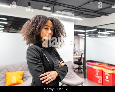 Selbstbewusste afroamerikanische Mitarbeiterin in schwarzer Jacke mit Afro-Frisur steht in einem modernen Arbeitsplatz und schaut auf die Kamera Stockfoto