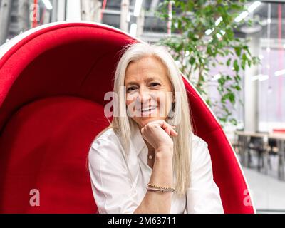 Positive ältere Geschäftsfrau, die lächelt und in die Kamera schaut, während sie sich während der Pause in einem modernen Arbeitsplatz auf einem aufgehängten roten Stuhl ruht Stockfoto