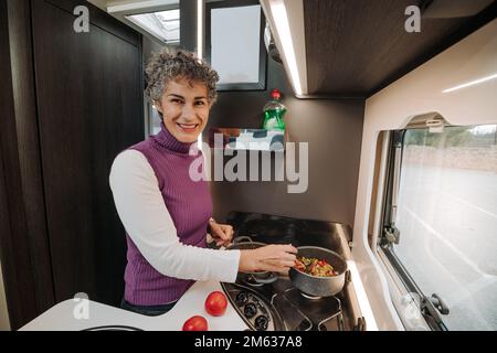 Positiv lächelnde Frau in lässiger Kleidung rührt Essen im Topf, während sie Abendessen im Wohnwagen kocht und auf die Kamera schaut Stockfoto