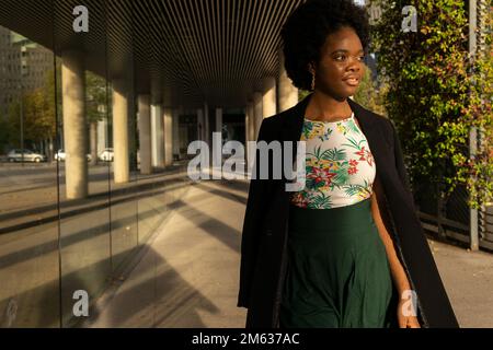 Nachdenkliche junge afroamerikanische Frau in schwarzer Jacke, die neben dem Gebäude steht und an sonnigen Tagen mit Bedacht wegschaut Stockfoto