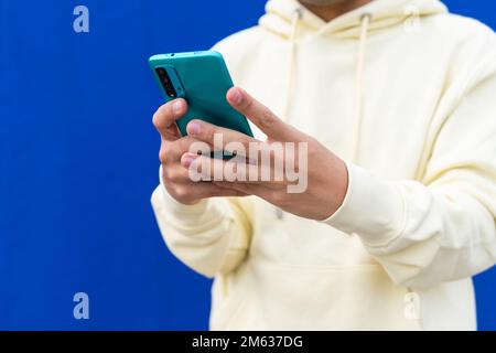 Von unten gekürzt, nicht wiedererkennbarer junger Mann in Kapuzenpullover, der auf dem Handy Textnachrichten schrieb, während er an der gelben Mauer der Stadtstraße stand Stockfoto