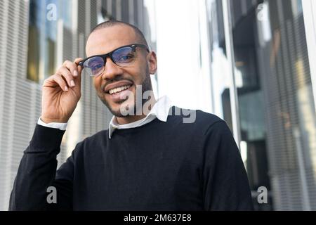 Ein Mann mit einem kleinen, lächelnden, ethnischen Bart in Brille und eleganter Freizeitkleidung, der neben dem Fenster des modernen Business Centers steht und in die Kamera schaut Stockfoto