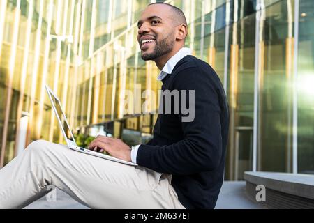 Seitenansicht eines lächelnden schwarzen Mannes in eleganter Freizeitkleidung, der Informationen auf einem Netbook eingibt, während er an einem geschäftlichen Projekt im Freien arbeitet Stockfoto