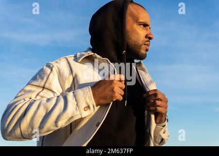 Ein männlicher Afroamerikaner in Jacke und Kapuze mit Kapuze auf dem Kopf, der auf der Straße gegen Sonnenlicht läuft und wegschaut Stockfoto