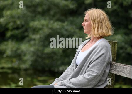 Seitenansicht einer aufmerksamen Frau mittleren Alters in lässiger Kleidung, die auf einer alten Holzbank neben einem Teich mit Lotusblättern saß Stockfoto