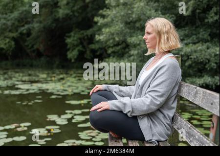 Ganzkörper-Seitenansicht einer Frau mittleren Alters in legerer Kleidung, die sich ausruht, während sie auf einer alten Holzbank in der Nähe eines Teichs mit Lotusblättern sitzt Stockfoto