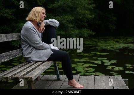 Ganzkörper-Seitenansicht einer Frau mittleren Alters in lässiger Kleidung, die auf dem Knie ruht, während sie auf einer alten Holzbank in der Nähe eines Teiches mit Lotusblättern sitzt Stockfoto