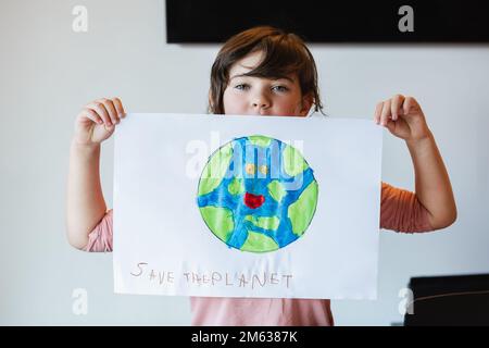 Ein Mädchen in legerer Kleidung, das Papier mit der Inschrift Erde und Save the Planet vorführt und im Klassenzimmer in die Kamera schaut Stockfoto