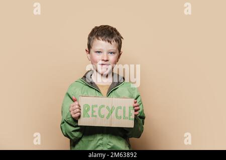 Ein Junge in grüner Oberbekleidung hält ein Recycle-Poster und schaut während des Umweltprotests vor beigefarbenem Hintergrund in die Kamera Stockfoto