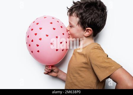 Seitenansicht eines niedlichen Jungen im braunen T-Shirt, der die Hand auf der Taille hält und einen rosa Ballon küsst, mit kleinen Herzen auf weißem Hintergrund Stockfoto