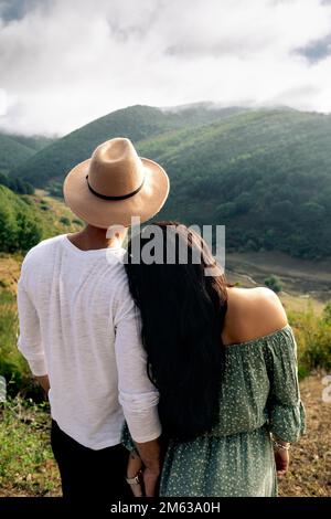 Rückansicht eines jungen weiblichen Mannes in romantischem Kleid und eines Mannes in legerem Outfit und Strohhut, der Händchen hält, während er die Berge genießt Stockfoto