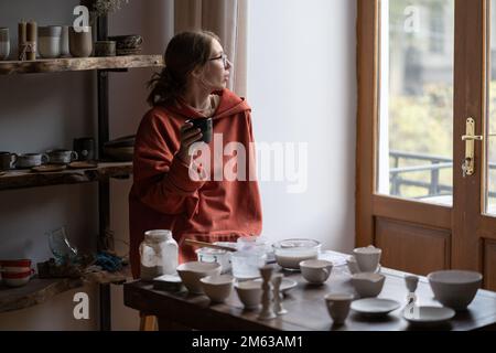 Träumige, pensive Töpferin, die eine Tasse Tee hält und aus dem Fenster blickt, während sie im Töpferstudio sitzt Stockfoto