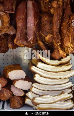 Geräucherte Fleischspezialitäten in verschiedenen Farben und Größen werden auf der Theke gestapelt. Fairer Handel mit hausgemachten Fleischspezialitäten. Nahaufnahme. Selektiver Fokus. Stockfoto