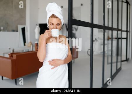 Porträt einer jungen Frau, die in ein Handtuch gewickelt ist und heißen Tee oder Kaffee trinkt Stockfoto
