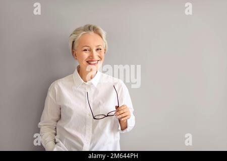 Glückliche, reife Geschäftsfrau, die an der grauen Copy Space-Wand steht, eine Brille hält und lächelt Stockfoto