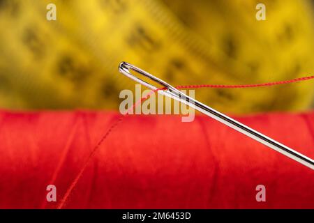 Nadelauge mit rotem Polyesterfaden. Spule aus rotem Polyester-Nähgarn. Maßband im Hintergrund. Stockfoto