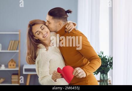 Ein glücklicher Mann umarmt seine Freundin, küsst sie auf die Wange und gibt ihr eine rote Valentinskarte Stockfoto