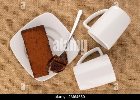Ein Stück Trüffelkuchen mit weißer Keramikuntertasse und zwei Tassen Jutetuch, Makro, Draufsicht. Stockfoto