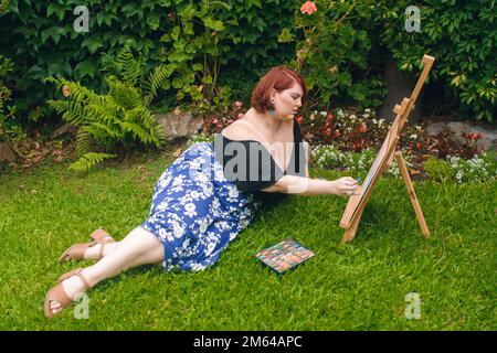 Rothaarige argentinische kaukasische Latina-Frau, plus Model, liegt auf dem Gras im Garten ihres Hauses, entspannende Zeichnung mit Kreide und Woo Stockfoto