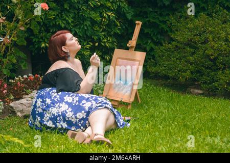 Junge weiße Latina-Frau, die auf dem Rasen mit einer Staffelei im Garten liegt, mit geschlossenen Augen denkt und atmet, während sie ihre Zeichnung beendet. Stockfoto