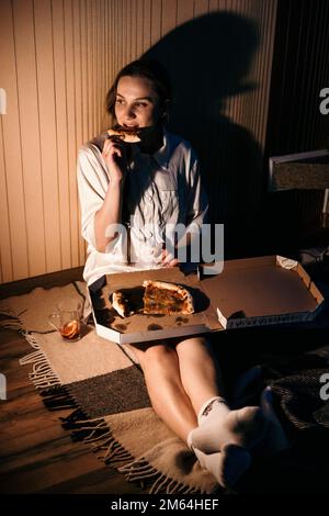 Einsamkeit in den Weihnachtsferien, Valentinstag. Junge, allein stehende Frau im Pyjama, die Pizza am Kamin im dunklen Haus isst. Stockfoto
