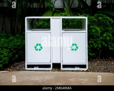 Grüne Recyclingsymbole auf modernen weißen, zweieisernen, rechteckigen Recyclingbehältern im grünen Garten vor dem Gebäude neben dem Betongang. Stockfoto