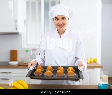 Fröhliche Chefköchin in weißer Uniform mit einem Backblech mit Muffins Stockfoto