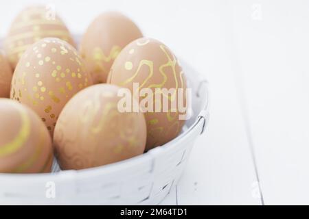 Braune Ostereier mit goldenem Muster in einem weißen Korb auf einem weißen Holztisch. Ostern Stillleben. 3D Rendern.. Stockfoto