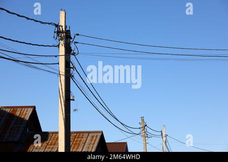 Powerline-Pfosten mit elektrischen Drähten und Kondensatoren über alten Dächern auf blauem Himmelshintergrund. Stromleitung, Stromversorgung im Dorf Stockfoto