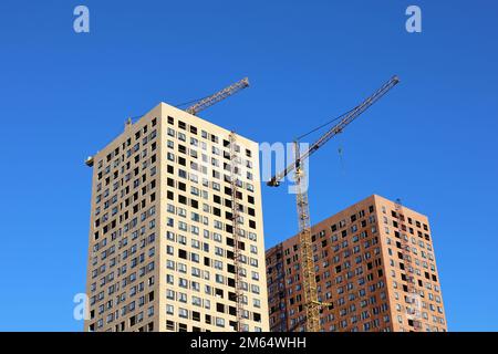 Turmkräne und zwei unfertige Gebäude vor blauem Himmel. Wohnungsbau, Wohnblocks in der Stadt Stockfoto