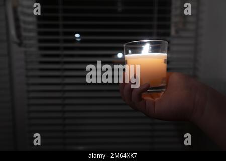 Das Mädchen hält eine Kerze in einem Glas in der Hand vor dem Hintergrund eines Fensters mit Jalousien (Nahaufnahme). Stromausfall. Energiekrise Stockfoto