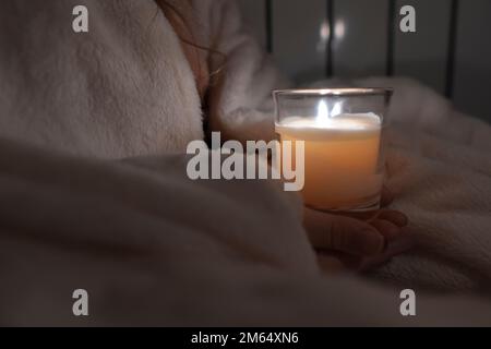 Ein Mädchen mit weißem Karomuster hält eine Kerze in einem Glasbecher in den Händen vor dem Hintergrund eines Heizkörpers (Nahaufnahme). Stromausfall. Energiekrise Stockfoto