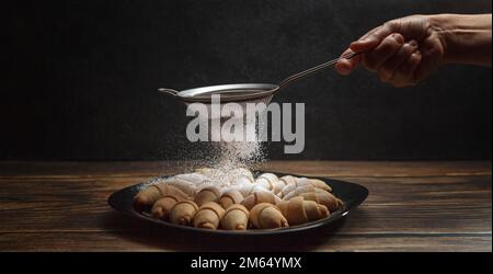 Frauenhände streuen Puderzucker durch ein Sieb auf frisch gebackene französische Croissants. Hausgemachtes Backkonzept Stockfoto