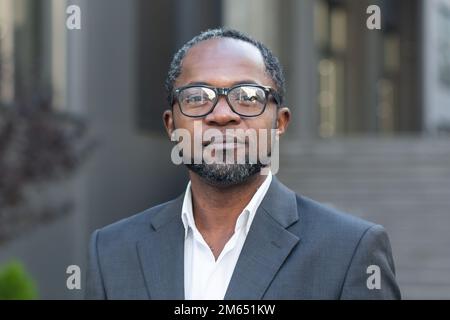 Nahaufnahme eines seriösen Geschäftsmanns, afro-amerikanischer Mann in Business-Anzug und Brille, der in Kameradenken schaut, Boss außerhalb des Bürogebäudes. Stockfoto