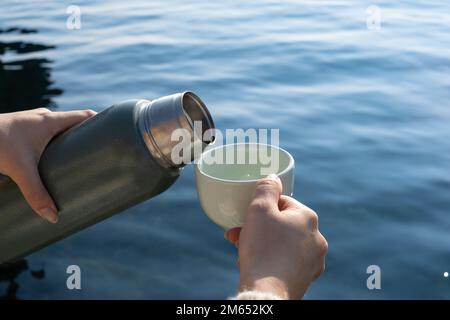 Die Hand einer Frau schüttet heißes Getränk aus der Thermoskanne in das Glas. Beim Gießen eines Getränks aus einer Thermoskanne in ein Glas ist das Meer im Hintergrund. Stockfoto