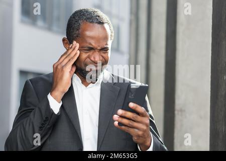 Ein afroamerikanischer Chef außerhalb des Bürogebäudes mit einem Telefon in den Händen, der online schlechte Nachrichten las, erhielt eine Benachrichtigung über Geldverlust und Betrug, verärgerte Geschäftsleute. Stockfoto