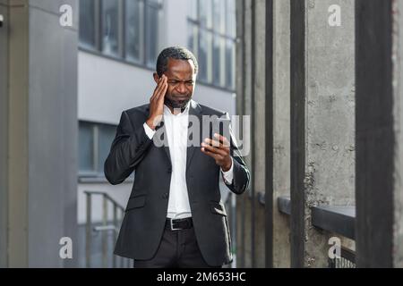 Ein afroamerikanischer Chef außerhalb des Bürogebäudes mit einem Telefon in den Händen, der online schlechte Nachrichten las, erhielt eine Benachrichtigung über Geldverlust und Betrug, verärgerte Geschäftsleute. Stockfoto