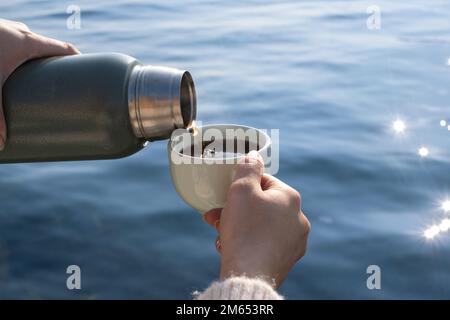 Die Hand einer Frau schüttet heißes Getränk aus der Thermoskanne in das Glas. Beim Gießen eines Getränks aus einer Thermoskanne in ein Glas ist das Meer im Hintergrund. Stockfoto