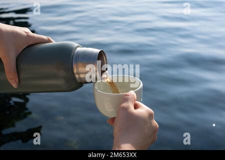Die Hand einer Frau schüttet heißes Getränk aus der Thermoskanne in das Glas. Beim Gießen eines Getränks aus einer Thermoskanne in ein Glas ist das Meer im Hintergrund. Stockfoto