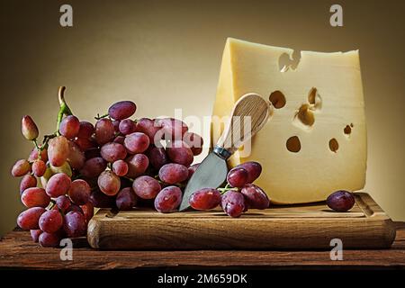 Großes Stück Käse mit Rosentraube und kleines Messer auf gelbem Hintergrund mit Holzbrett Stockfoto