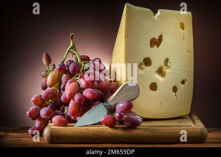 Ein großes Stück Käse mit Rosentraube und ein kleines Messer auf einem braunen, verlaufenen Holzbrett Stockfoto