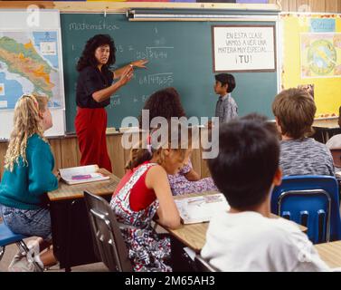 Hispanische Lehrerin, die an der Tafel steht, in einem Klassenzimmer voller Schüler im Grundschulalter, die sich mit Unterteilungsproblemen beschäftigen Stockfoto