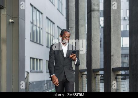 Glücklicher erfolgreicher afroamerikanischer Geschäftsmann, der vor dem Bürogebäude spaziert, mit dem Manager, der mit dem Smartphone Nachrichten liest und lächelt, mit der Kommunikations-App. Stockfoto