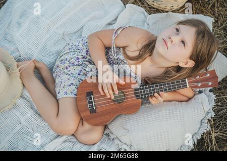 Nettes kleines Mädchen spielt Ukulele auf der Decke auf dem Feld. Porträt eines Kindes beim Aufschauen. Spaziergänge im Freien und Picknick. Hölzernes Musikinstrument, Hobby. S Stockfoto